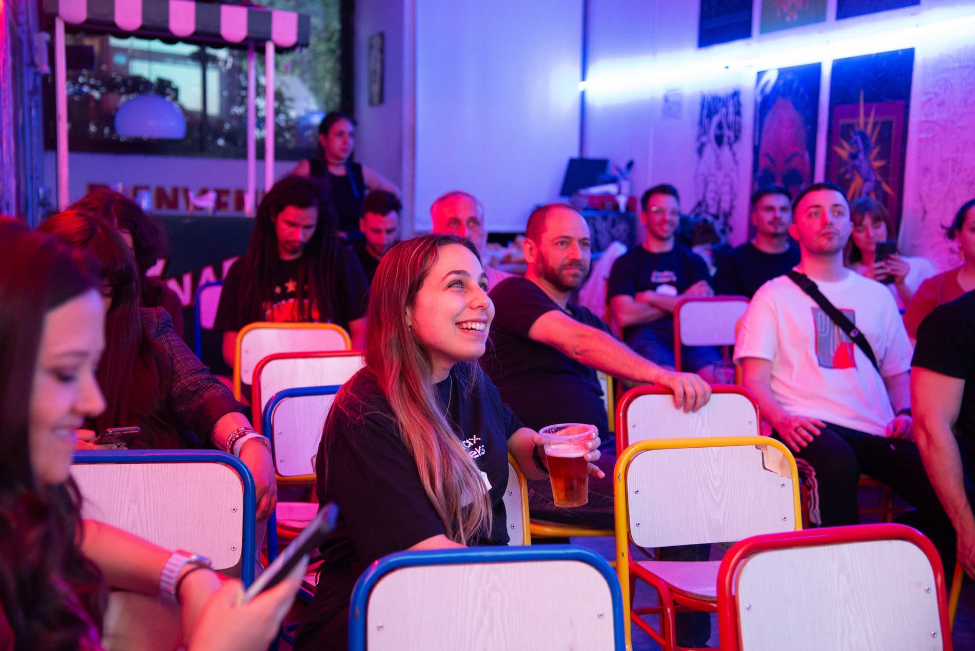 A group of people seated in a brightly lit room with colorful chairs at an event in Buenos Aires. A woman in the foreground is smiling and holding a drink, while others around her are engaged and listening. The room has a vibrant atmosphere with neon lighting and artwork on the walls.